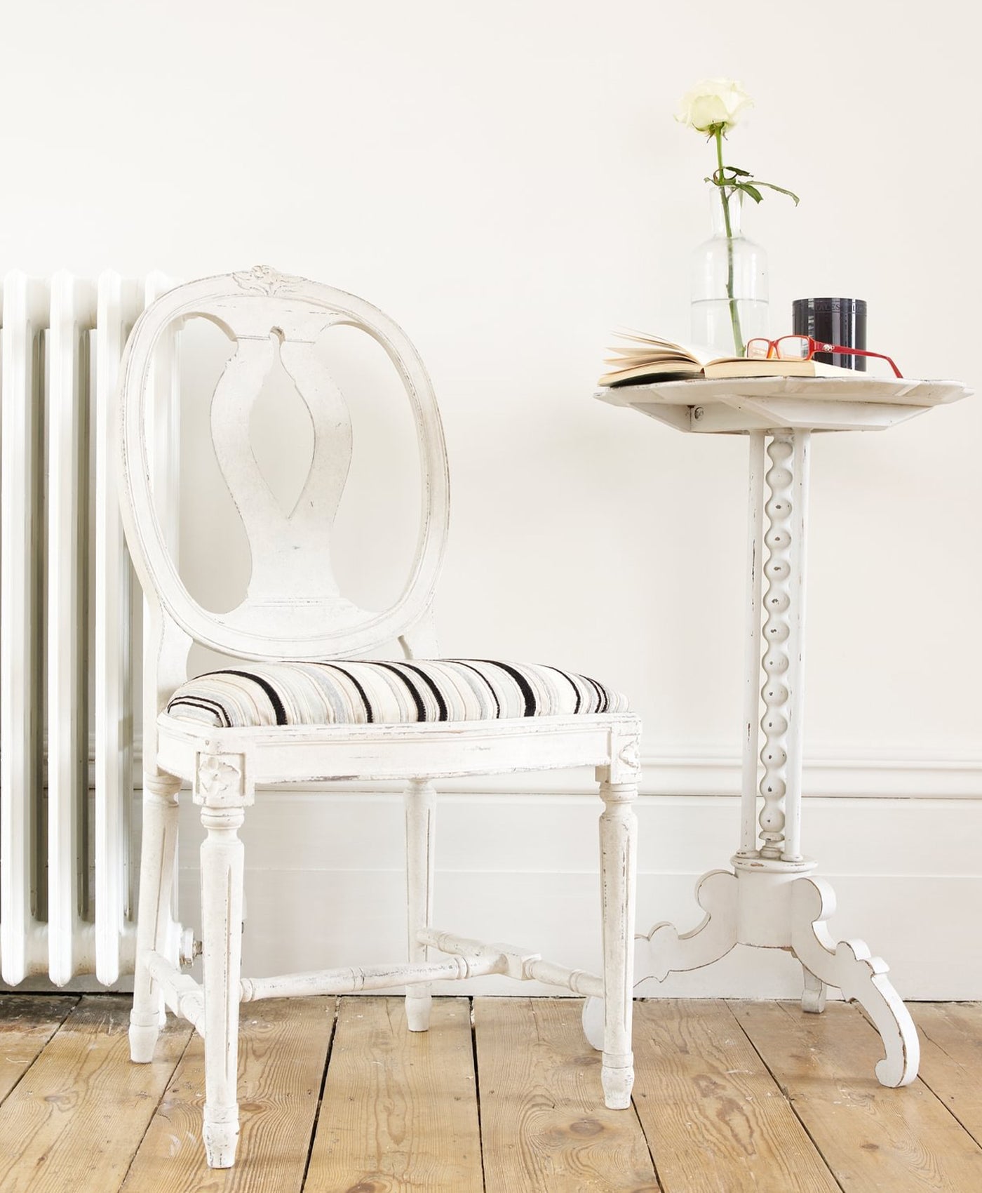 White chair with striped cushion next to a small table with a flower and books on a wooden floor.