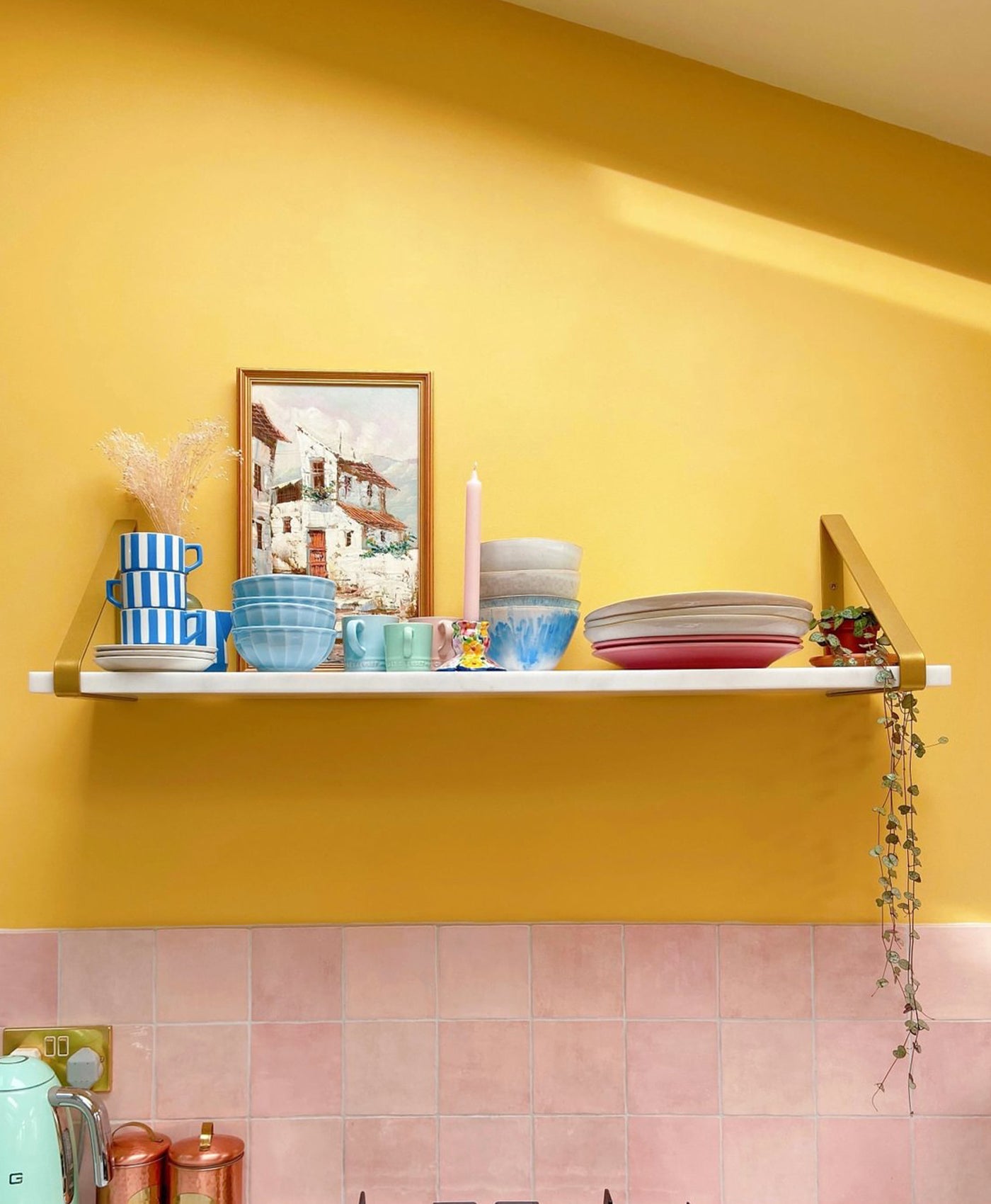 Shelf with colourful dishes against a vivid yellow wall with pink tiles.
