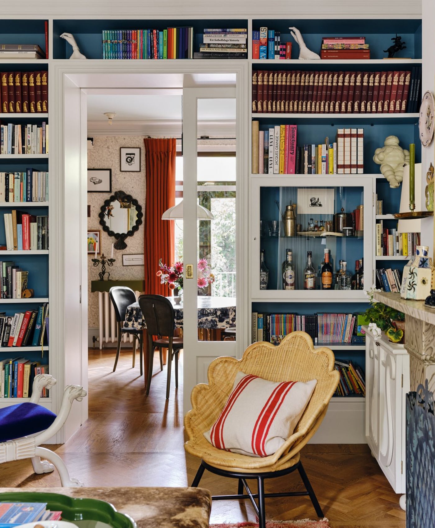 Beata Heuman's Living room with blue bookshelves, a wicker chair, and a view into another room.