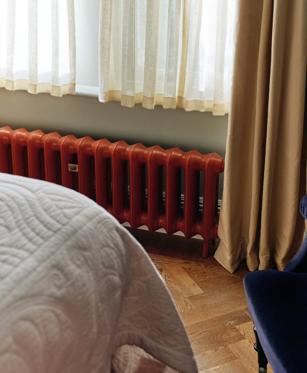 Red radiator against a wall with curtains and a bed in the foreground