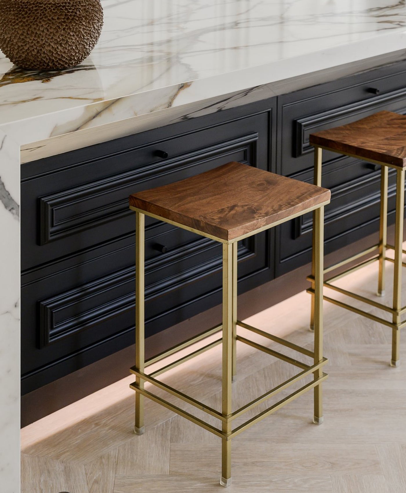 Two wooden stools with gold legs in front of a black kitchen island with marble countertop.