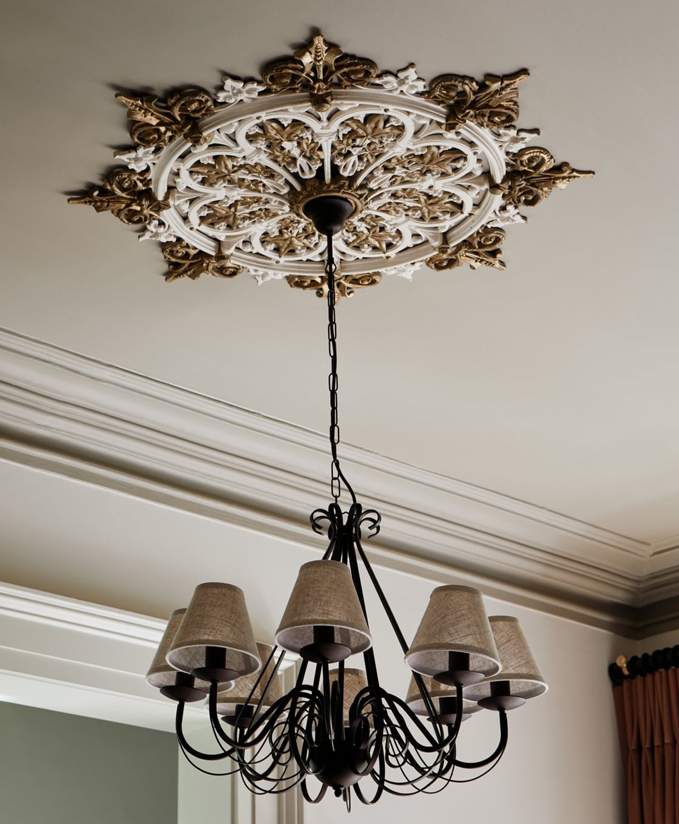 Decorative chandelier hanging from a ornate ceiling rose in a room with white walls and a window.