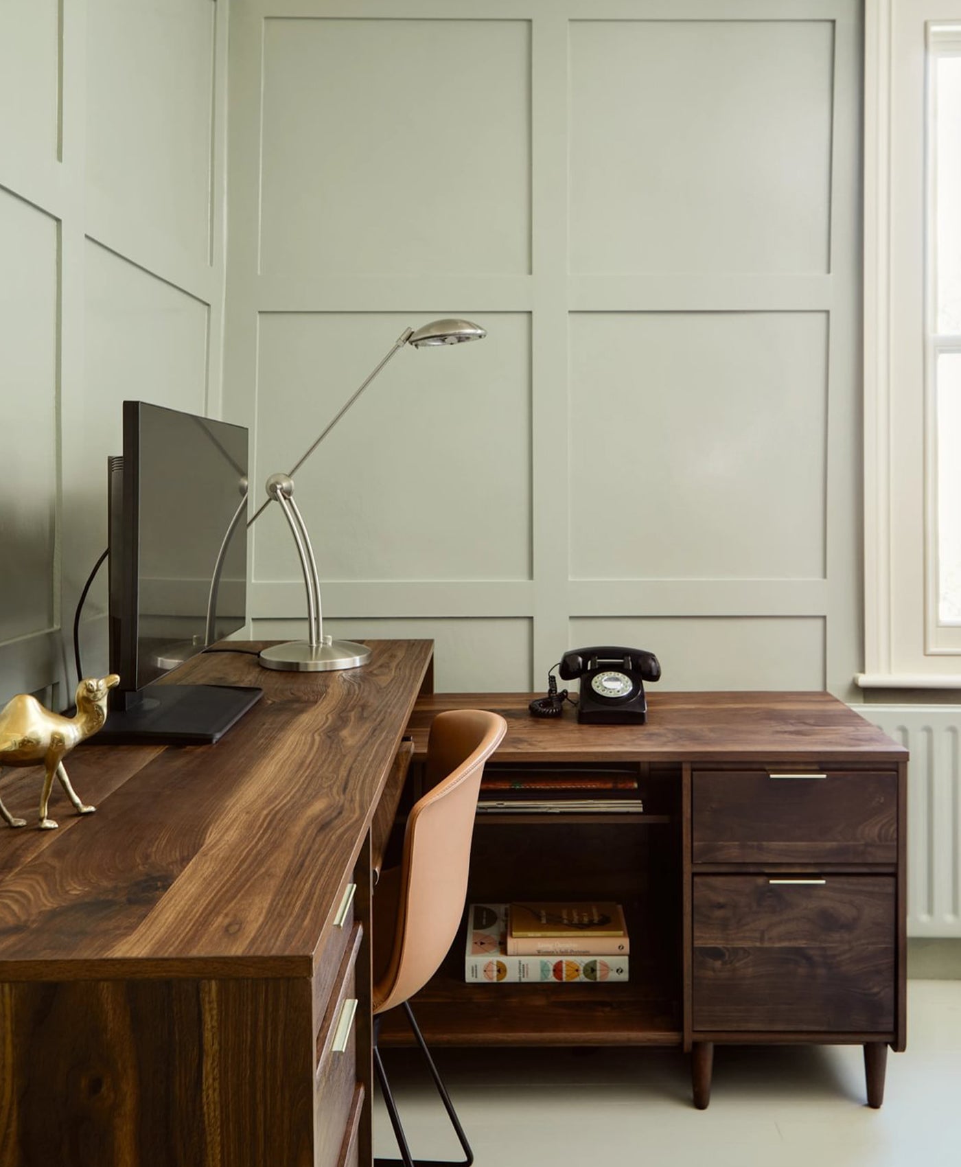 Wooden desk with a chair, lamp, and vintage phone in a home office setting with desaturated olive green wooden panelling on walls.