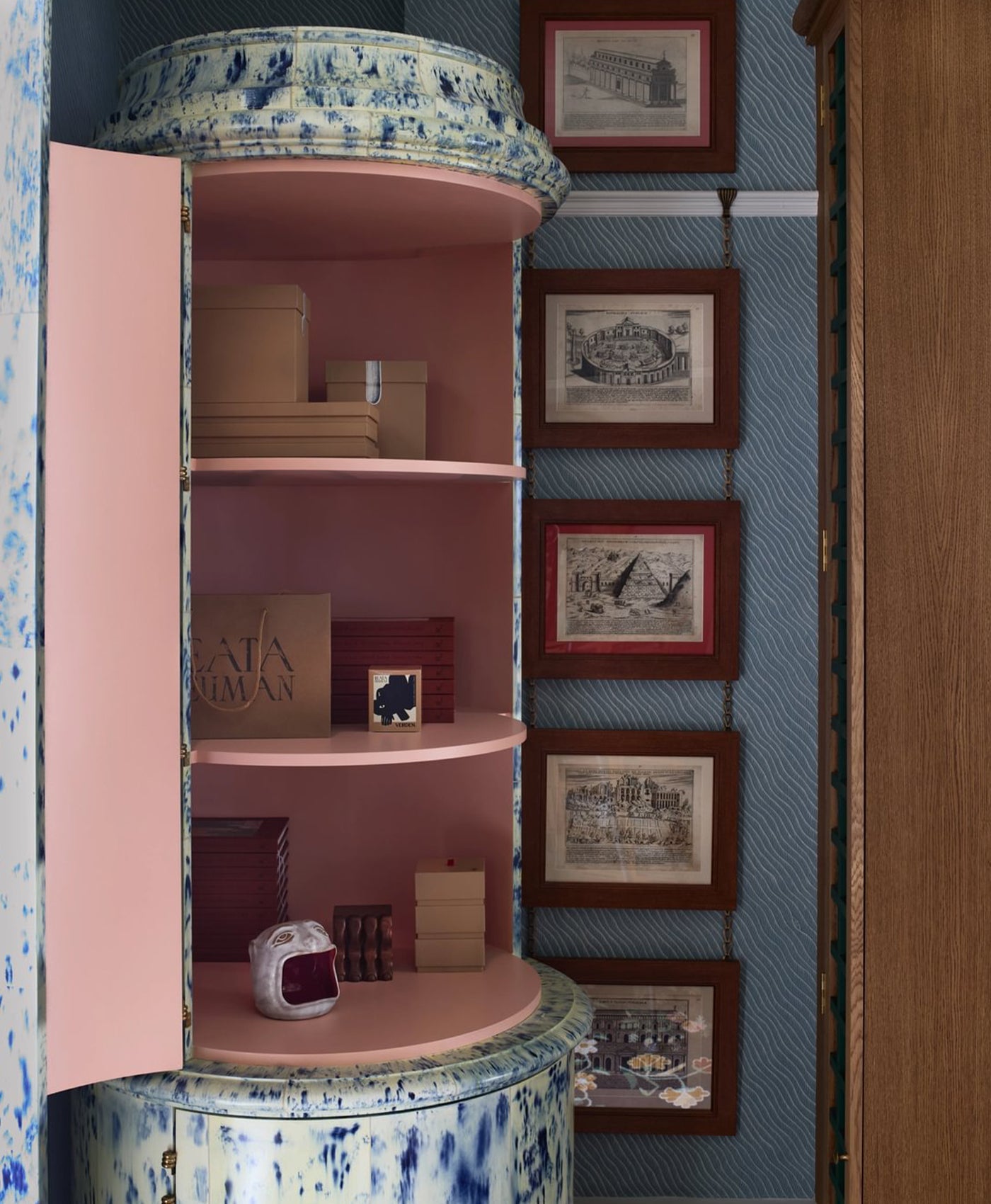 Decorative shelf with flesh pink interior against a blue patterned wall with framed pictures.