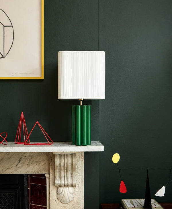 Green lamp with white shade on a marble surface against a dark green wall.