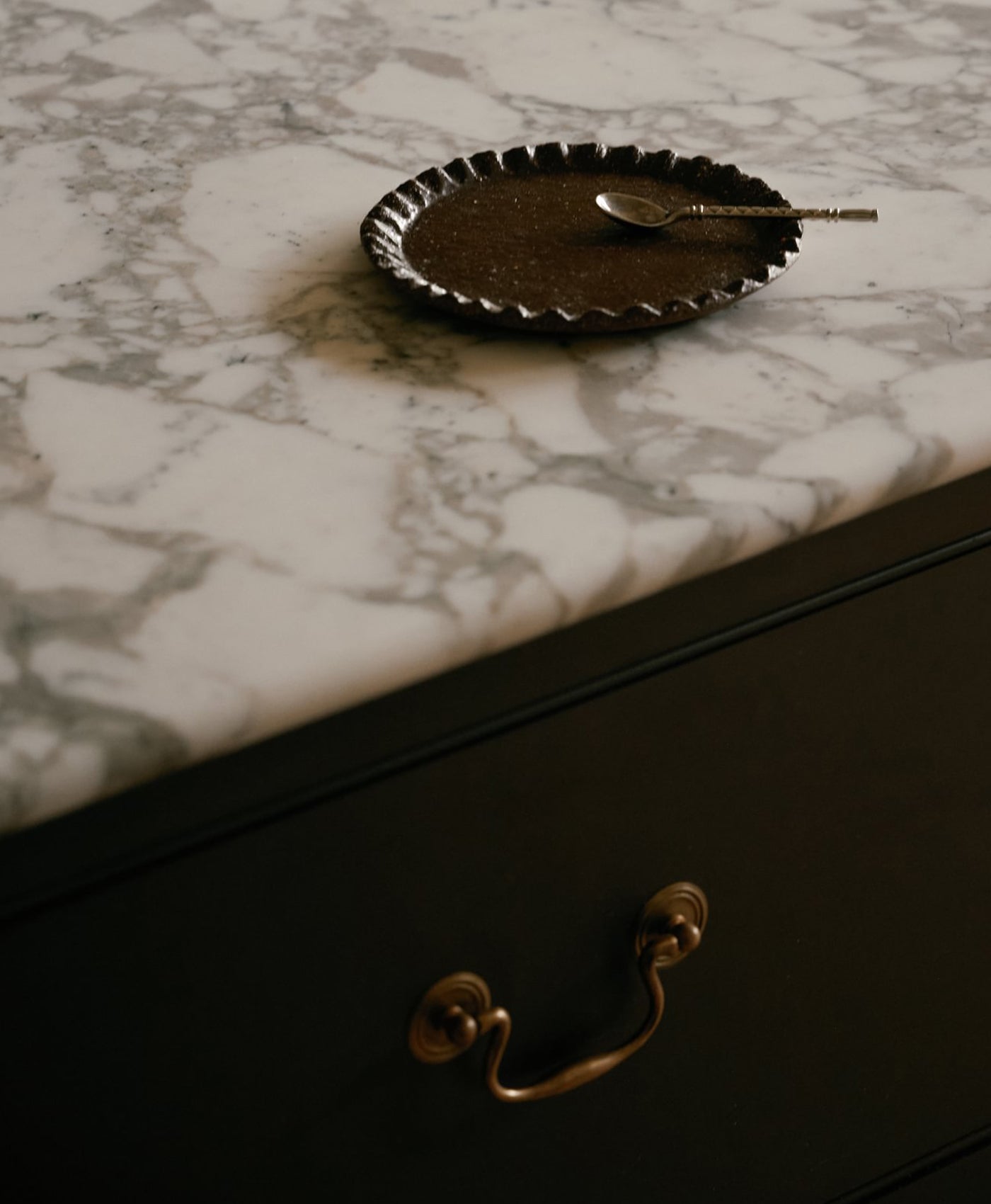Small round black dish with a spoon on a marble surface on top of a dark brown kitchen drawer