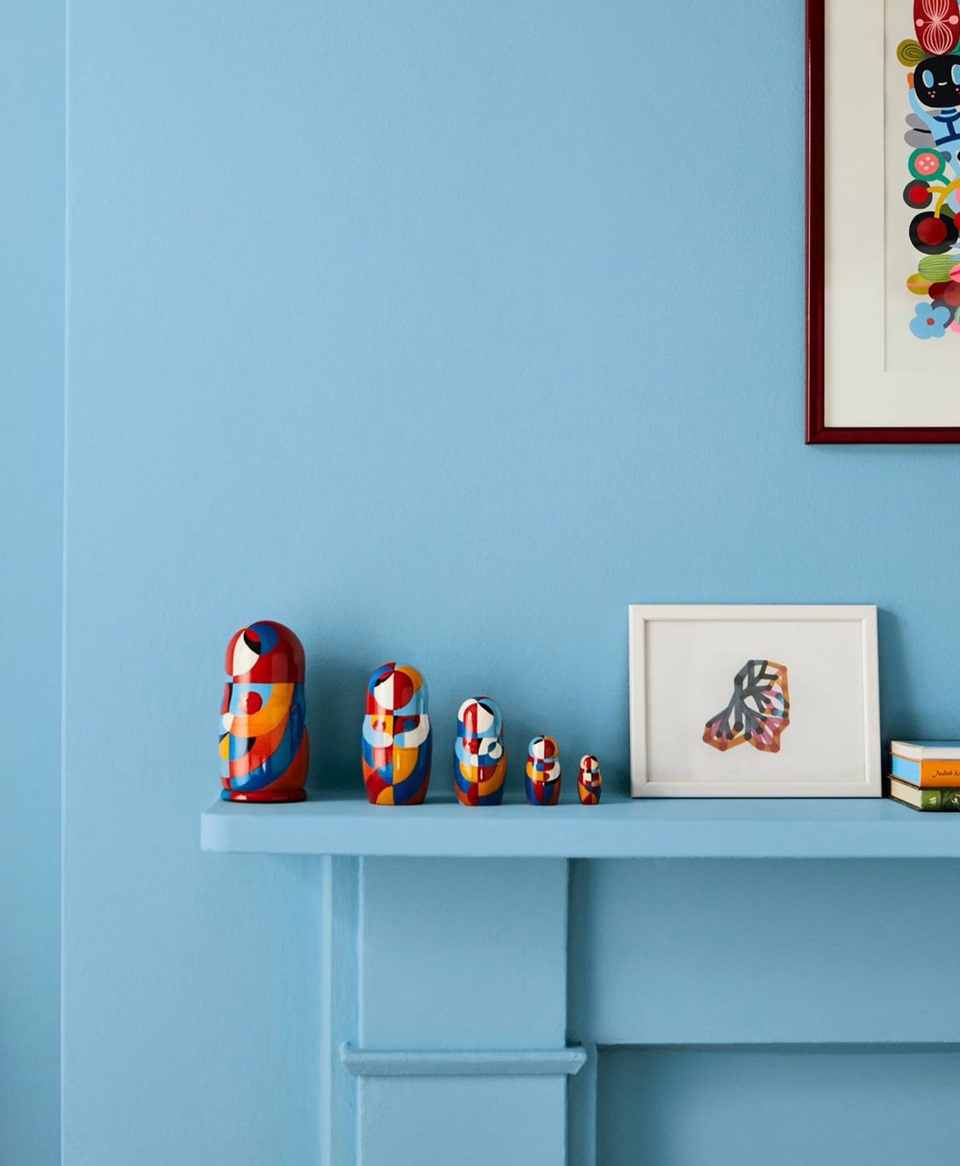 Colorful nesting dolls on a blue shelf against a light blue wall with framed artwork.