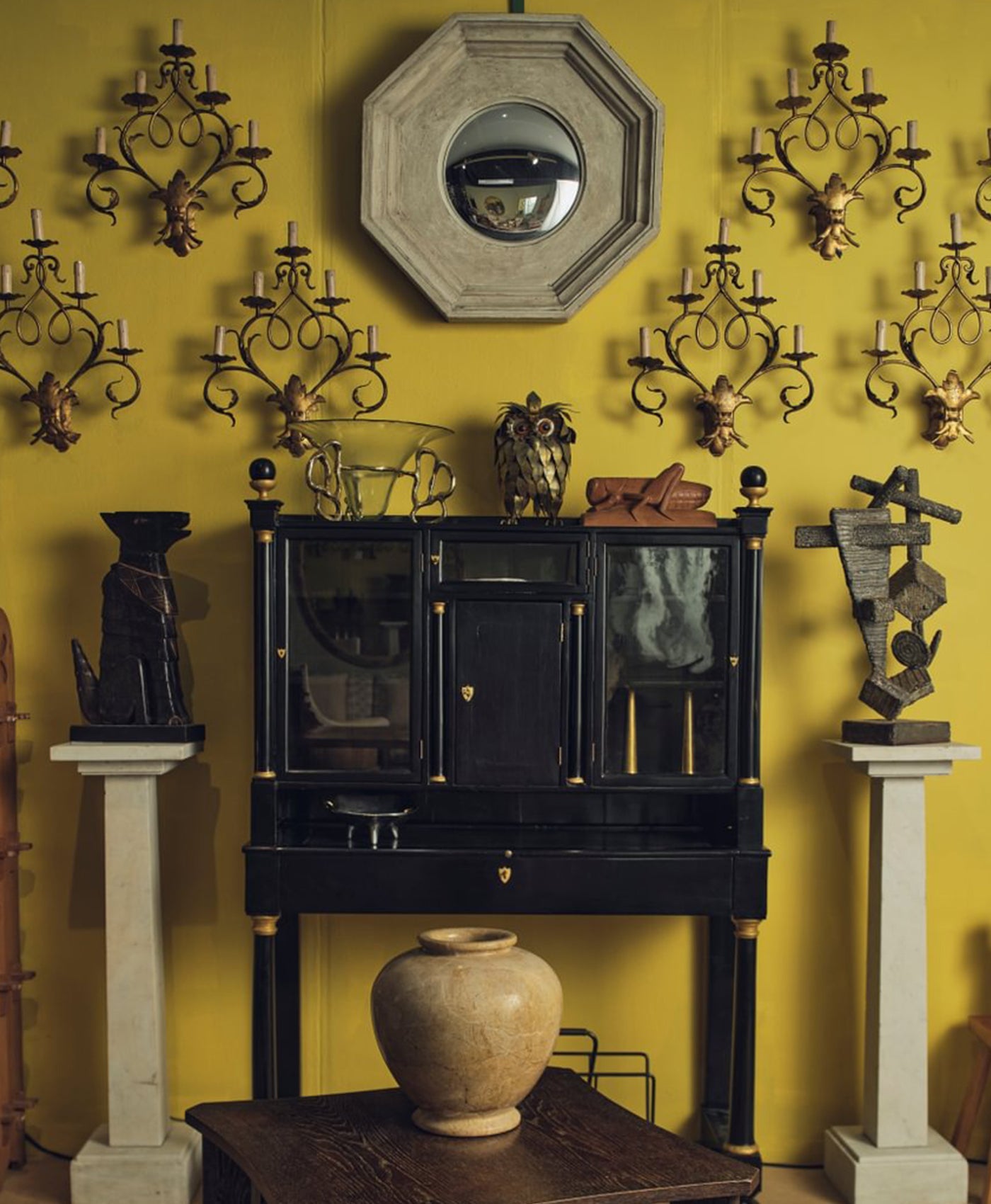 Decorative wall with a yellow background featuring a black cabinet, sculptures, and wall-mounted candle holders.