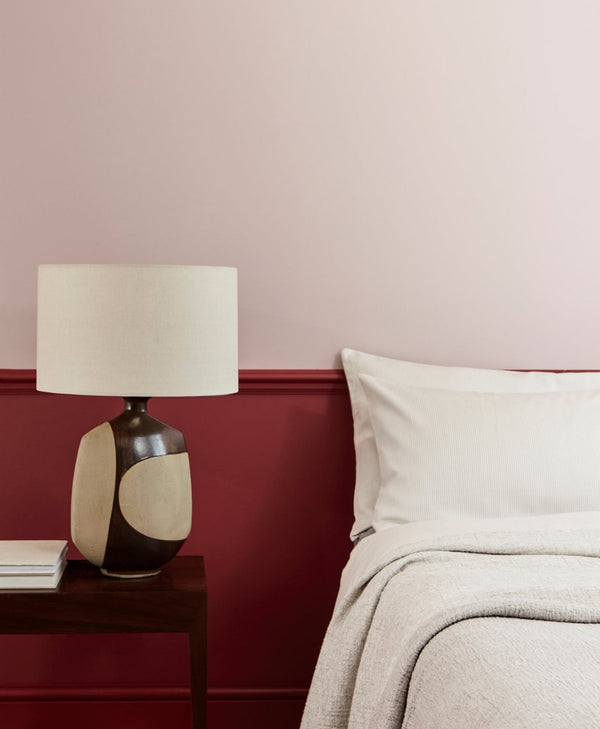 Decorative lamp on a nightstand next to a bed with a pale pink wall with deep red panelling