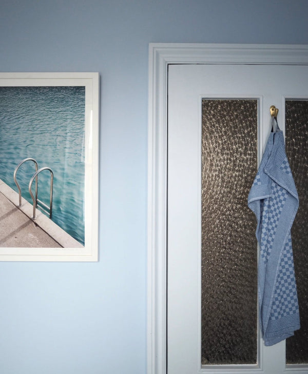 Light blue bathroom, Blue towel hanging on a door with a framed picture of a pool on the wall.