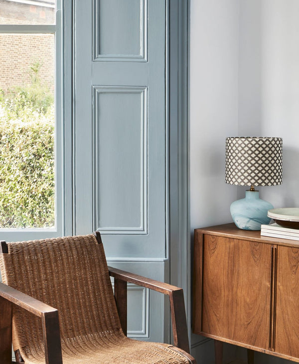 Wicker chair and wooden sideboard with lamp against a light blue wall and dusty blue wooden trims