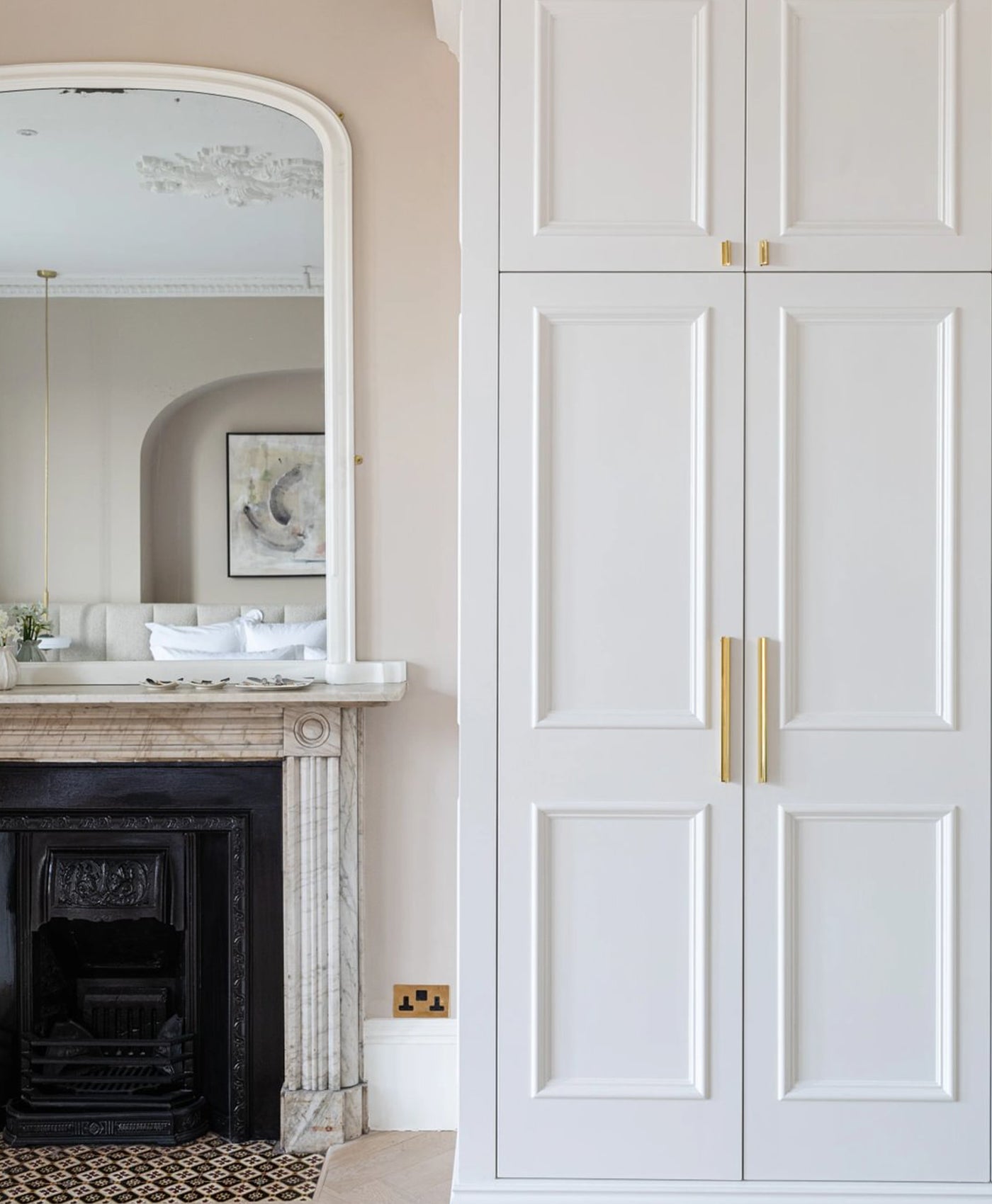Warm neutral bedroom, with white wardrobe with gold handles next to a fireplace with an arched mirror above.