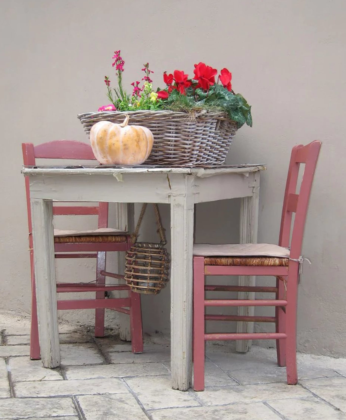 Vintage-style table with pink chairs and a basket of flowers against a plain wall.