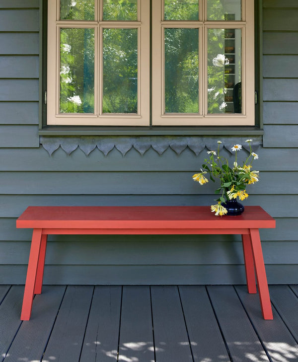 Red bench on a deck with a window and greenery in the background