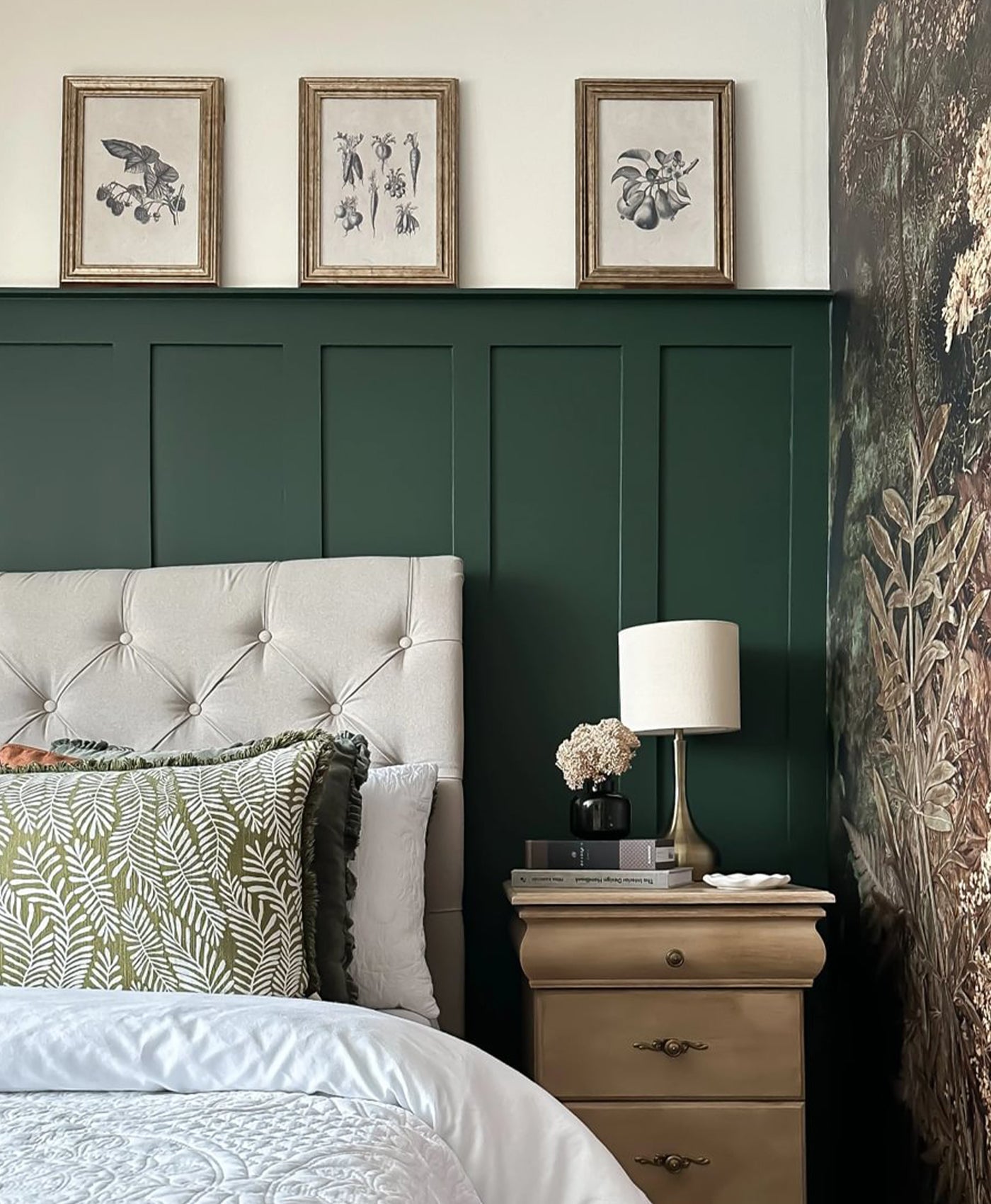 Neatly made bed with decorative pillows against a dark green panelled wall with framed artwork.