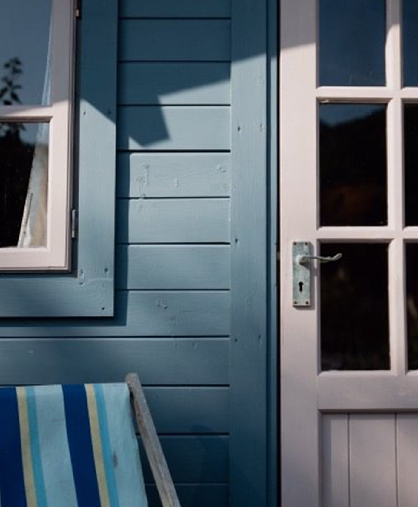 Blue wooden shed with a door and window, featuring a striped deck chair.