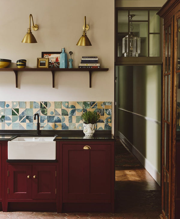 Kitchen interior with a colourful backsplash, wooden cabinets painted in a deep red, and shelves. Walls painted in plaster coloured paints.