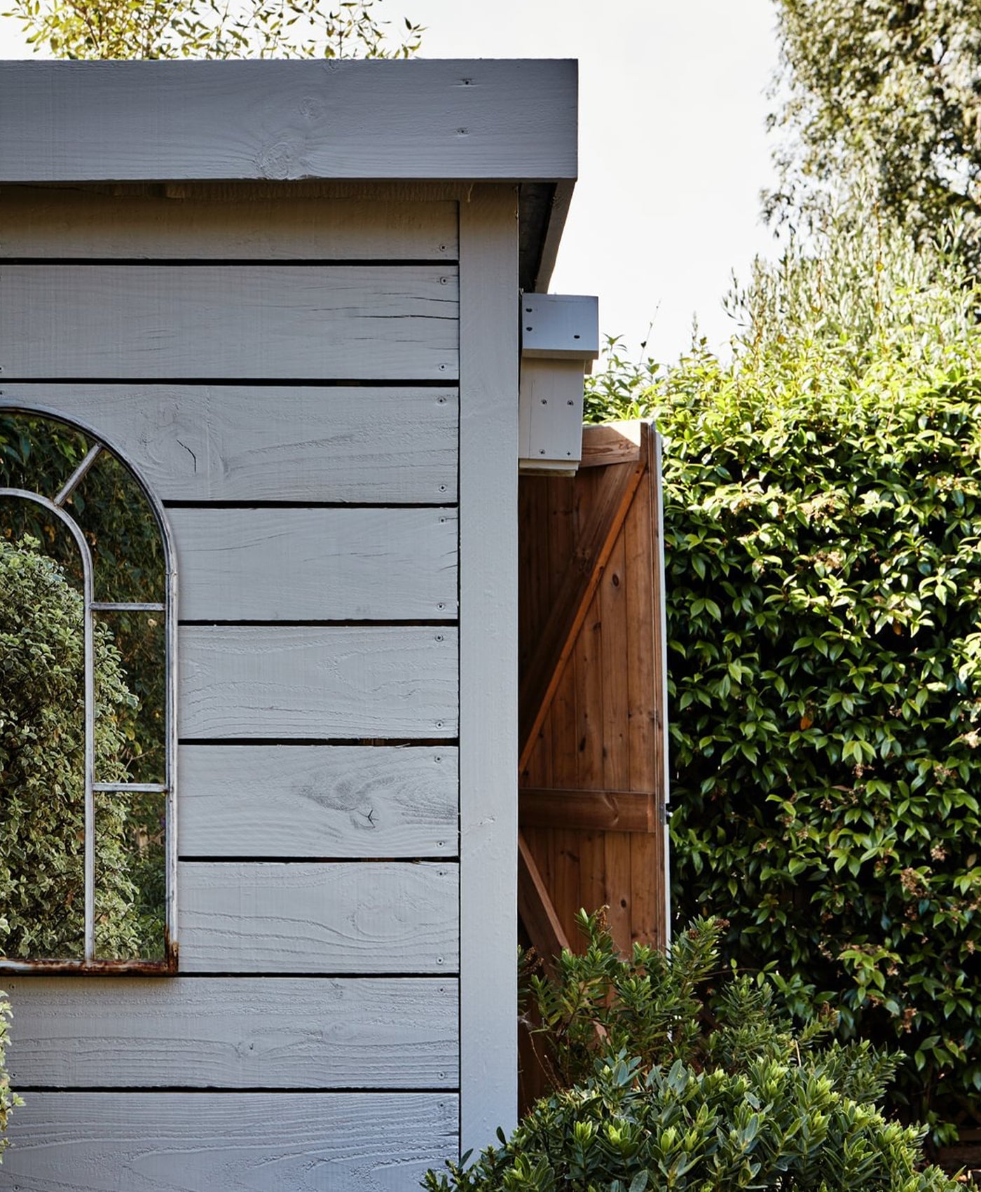 Wooden shed painted in grey paint with a slanted roof and arched window, surrounded by greenery.