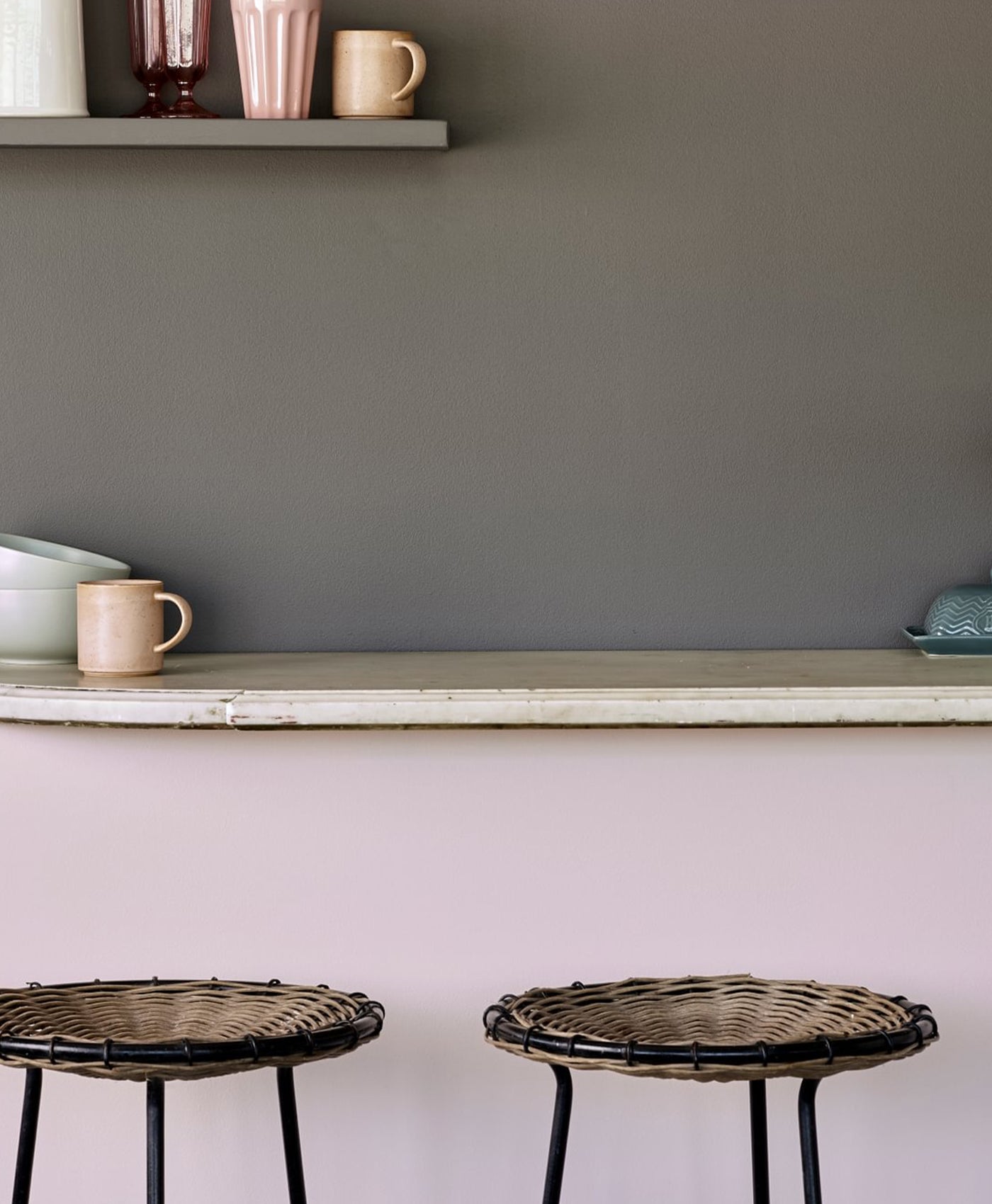 Two wicker stools in front of a kitchen counter with a grey wall and shelf in the background.