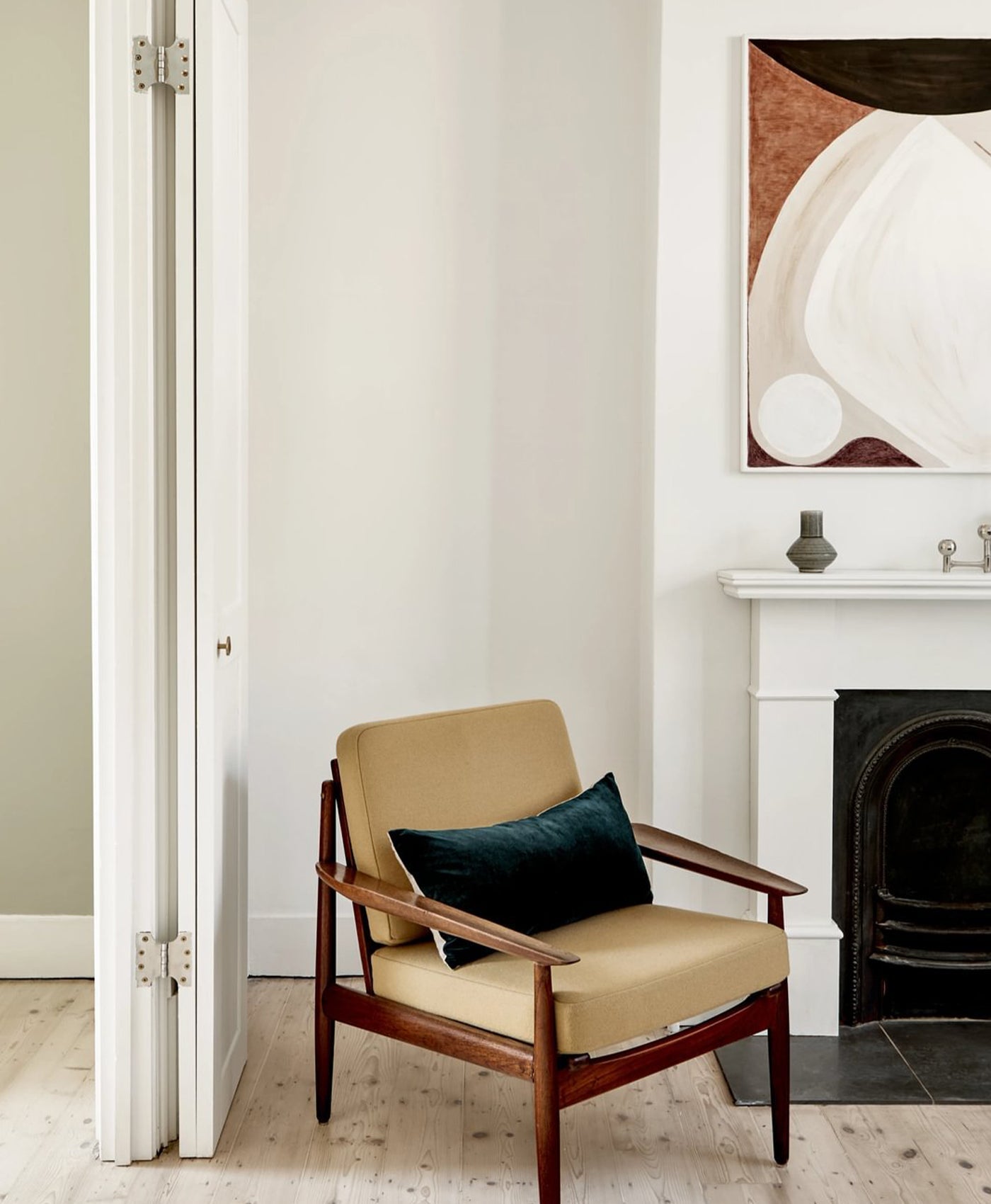 Beige armchair with a dark green pillow in a white room with a fireplace and abstract art.