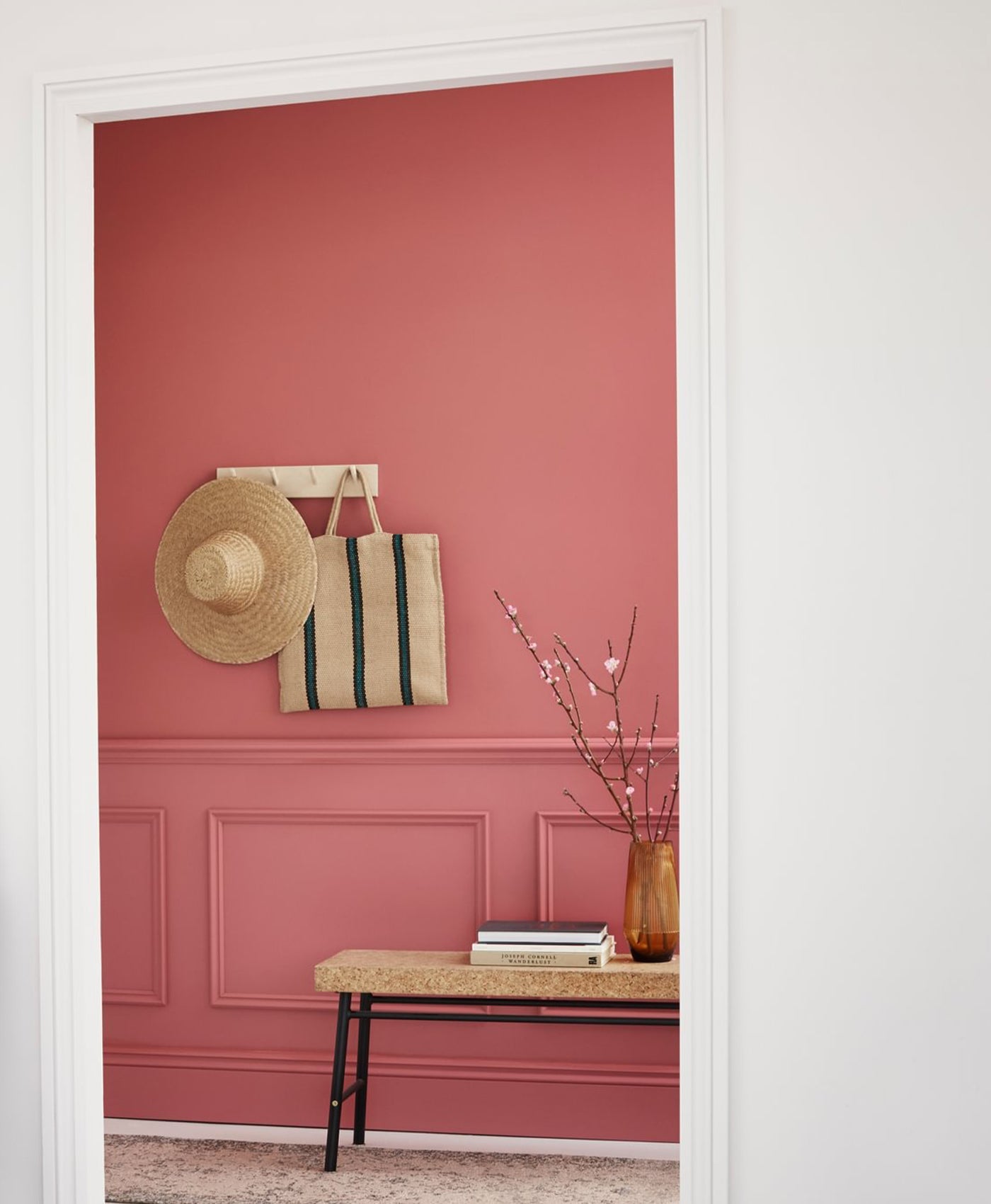 Red pink wall with a white frame, a table with a vase and books, and a hat and bag on a hook.