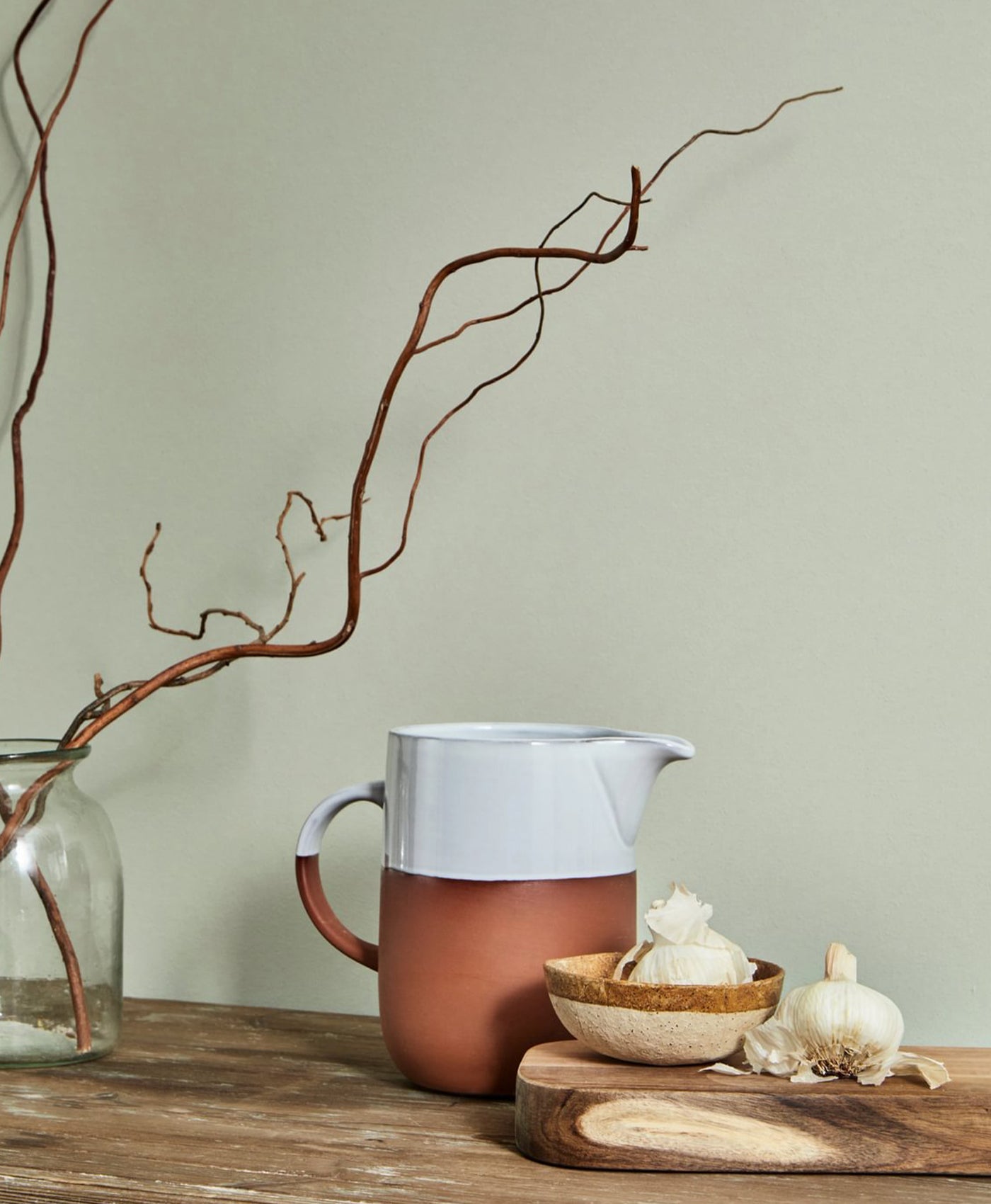Ceramic pitcher on a wooden surface with a branch and small items in the background, against light green wall