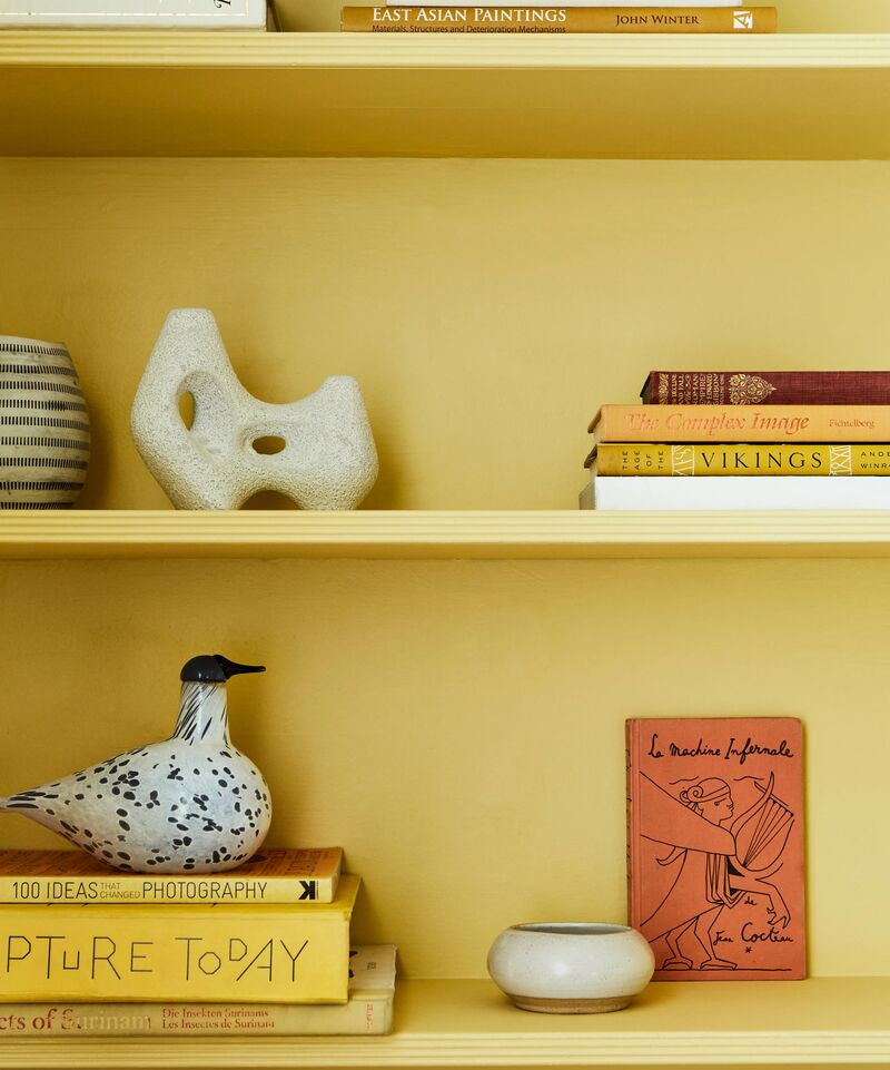 Bookshelf with decorative items including books and a ceramic bird on a yellow wall.
