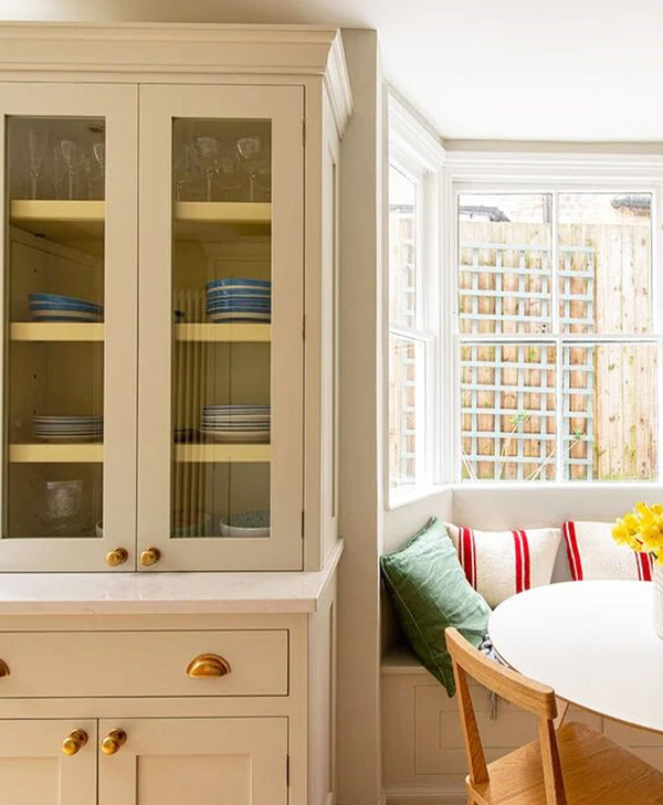 Beige kitchen cabinet with glass doors displaying plates, window seat with cushions, and a table.