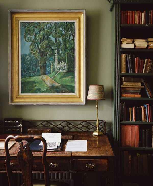 Framed painting on a pale olive green wall above a wooden desk with books and a lamp.