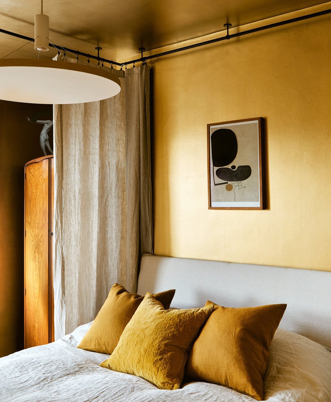 Bedroom painted in gold with mustard yellow pillows on a bed, framed artwork on the wall, and a wooden wardrobe.