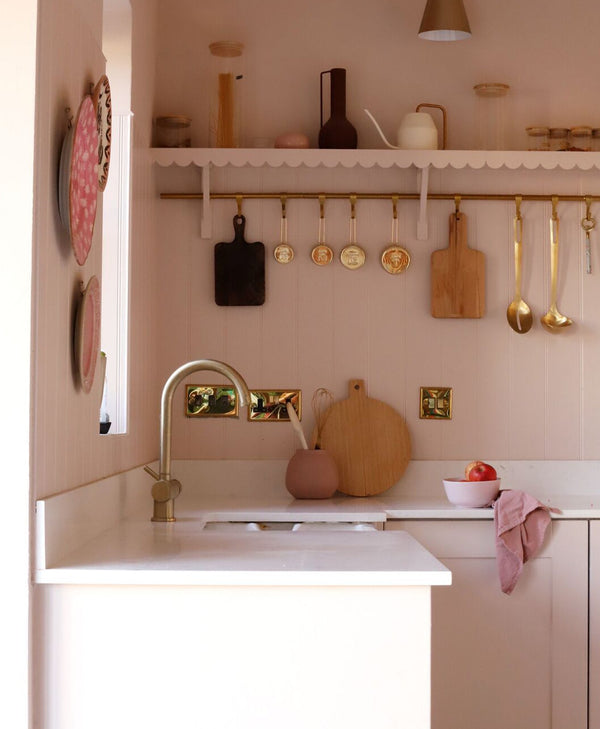 Modern pale pink kitchen with white countertops, gold fixtures, and various utensils hanging on the wall.