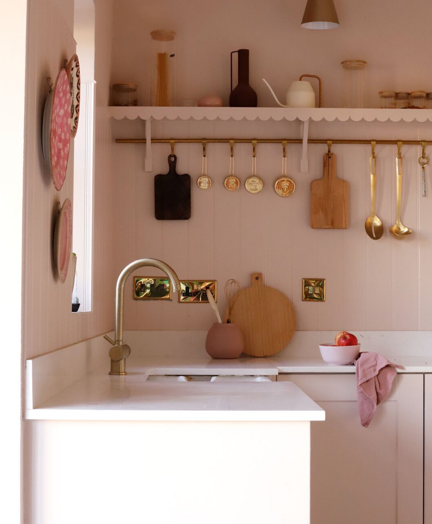Modern pale pink kitchen with white countertops, gold fixtures, and various utensils hanging on the wall.
