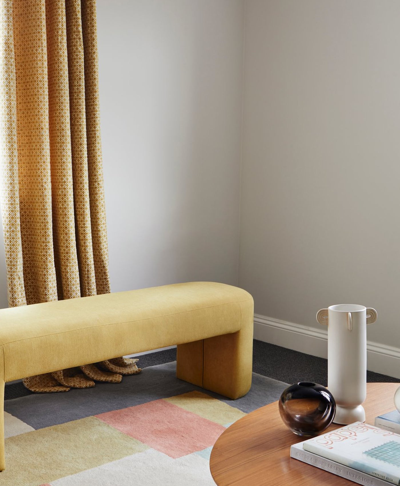 Yellow bench in a room with a warm grey wall, colourful rug, and a table with a vase and books.