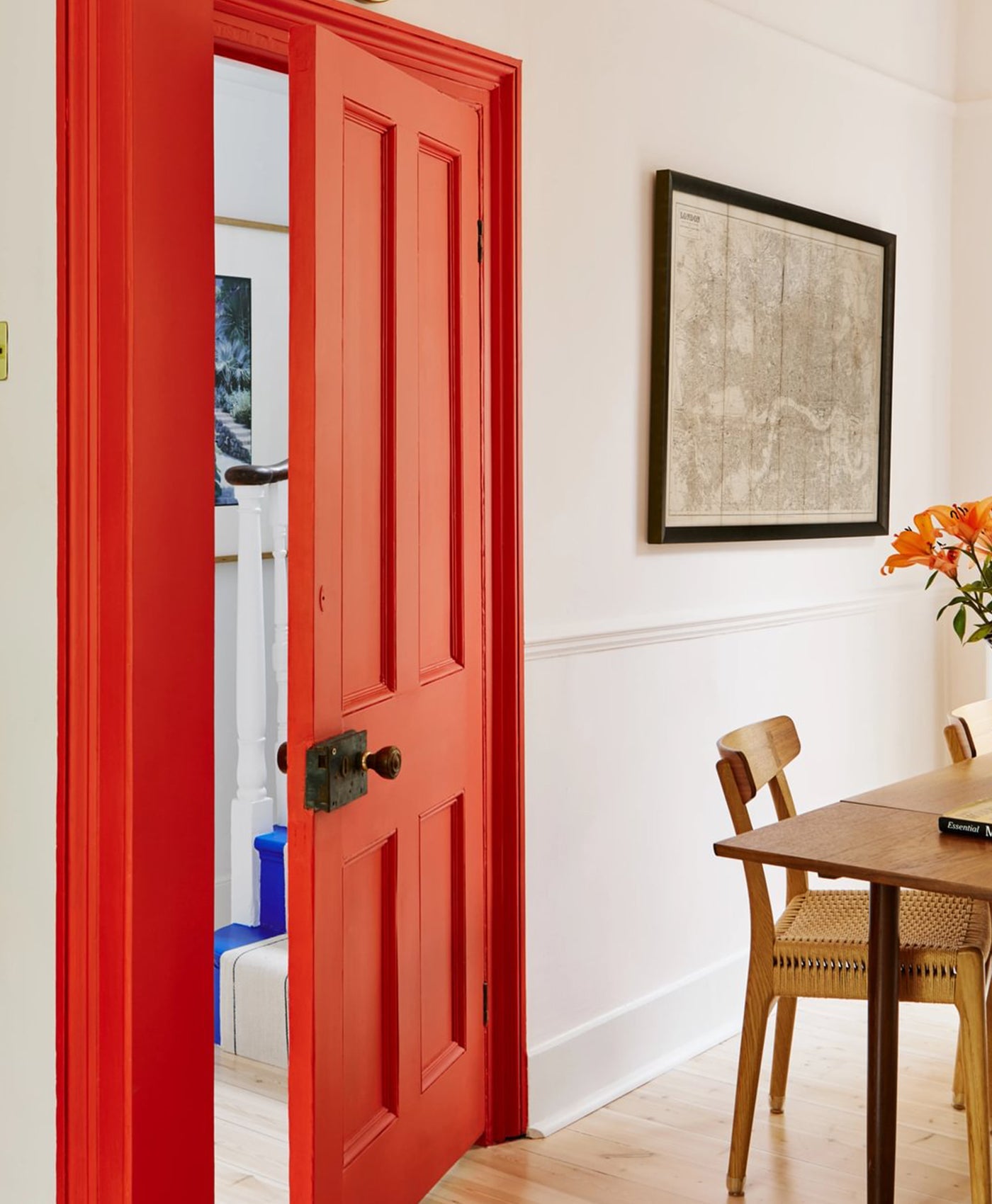 Pale pink room with red door open to a room with a wooden table and chairs, and a framed picture on the wall.