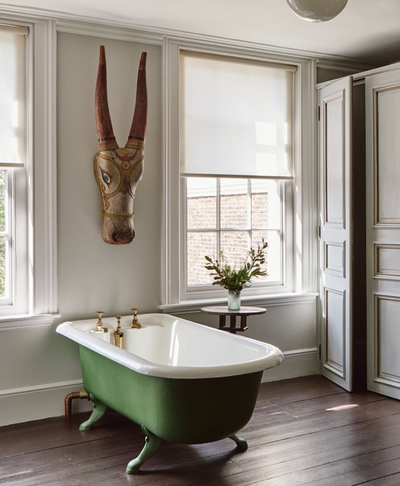 Green freestanding bathtub in a room with large windows and a decorative animal head on the wall. Wooden floor painted in dark brown paint.