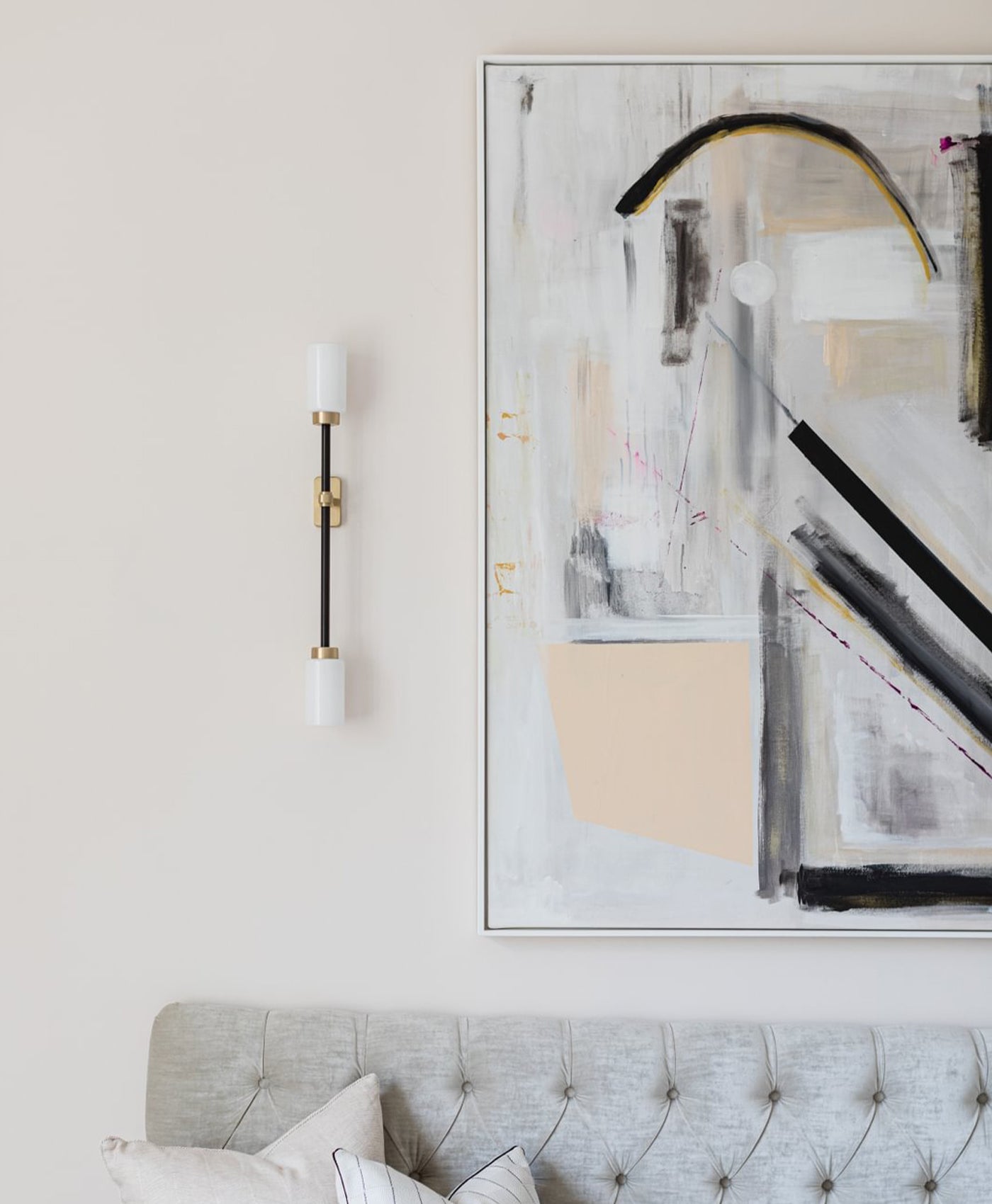 Abstract art piece on a wall above a grey sofa with a light pink wall and sconce in the background.