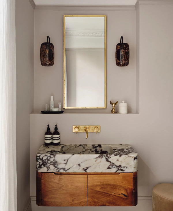 Bathroom vanity with marble countertop and wooden cabinet, featuring a gold faucet and mirror and walls painted in a pale plaster coloured paint