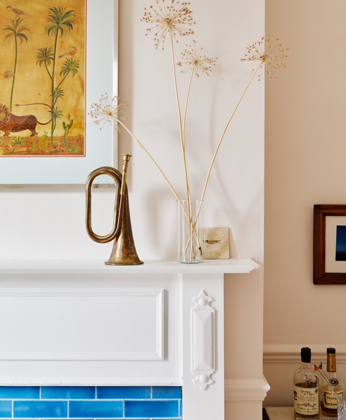 Decorative mantelpiece with a brass vase, glass vase with dried plants, and framed artwork.