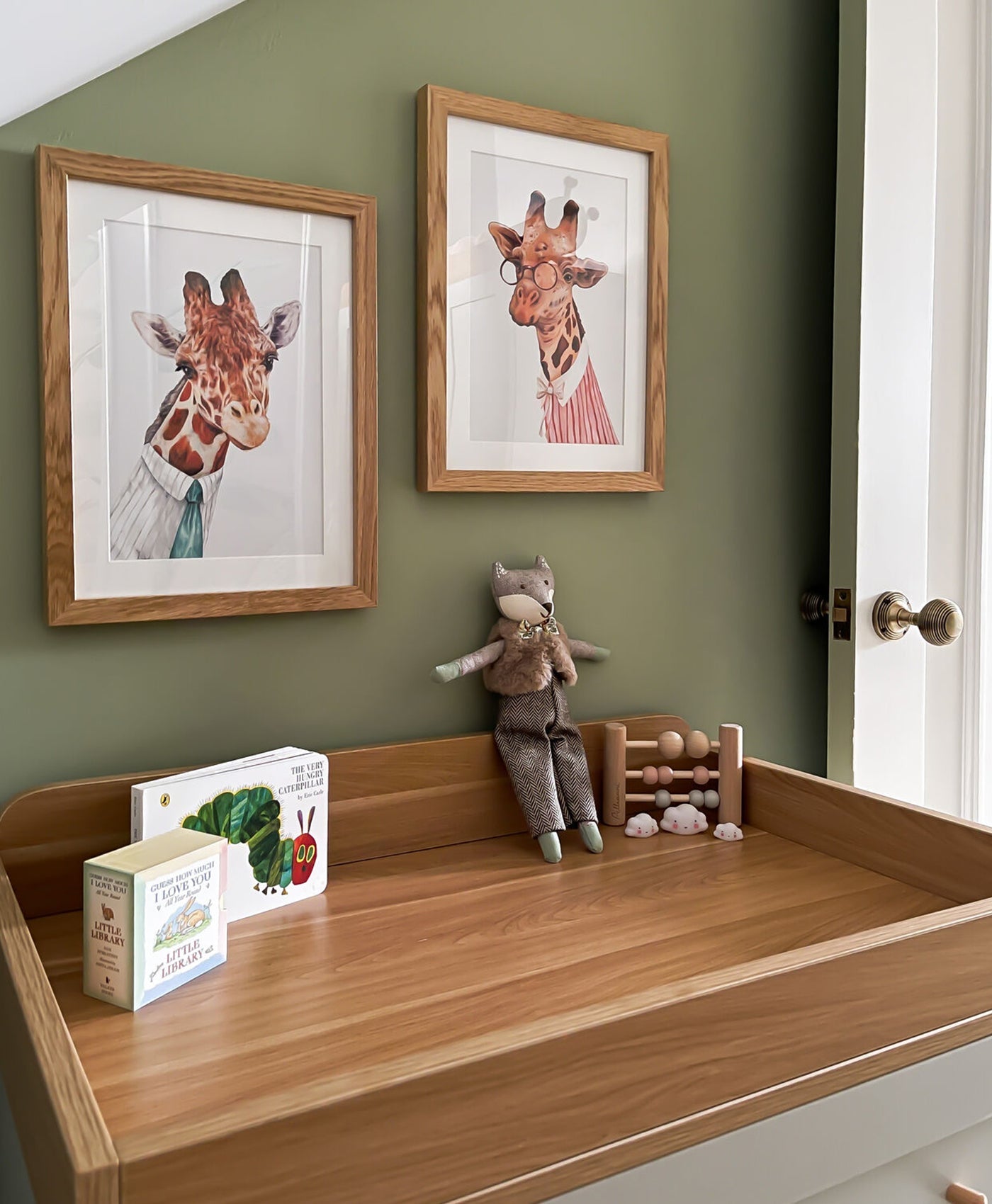 Wooden shelf in nursery with toys and framed pictures of giraffes against a grey green wall.