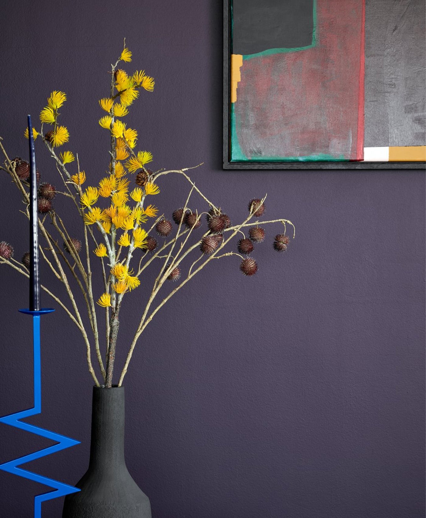 Decorative arrangement with yellow flowers and brown leaves in a gray vase against a purple wall with abstract art.