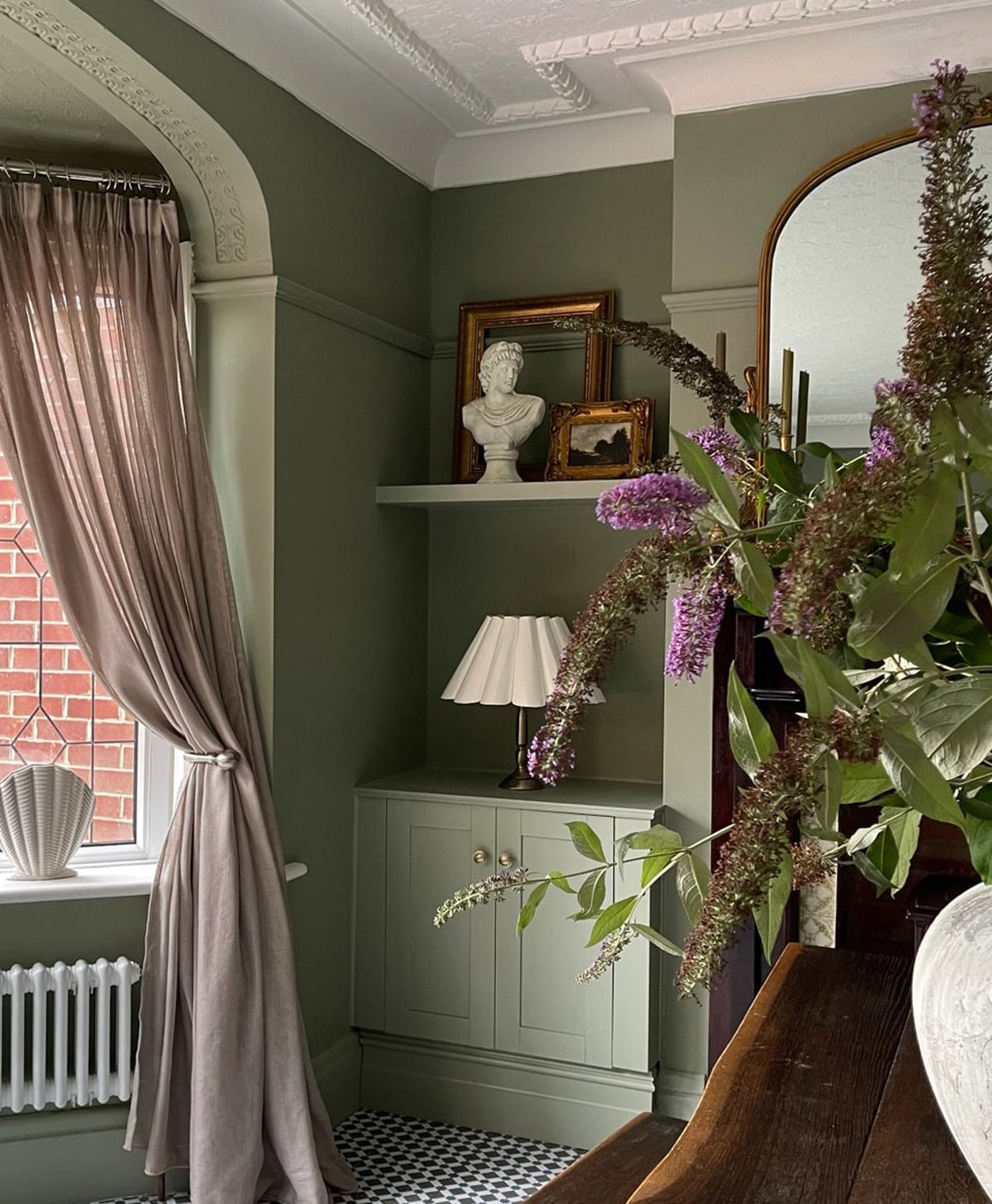 olive green victorian style living Room interior with a window, curtains, shelf with decor items, and a vase with flowers.