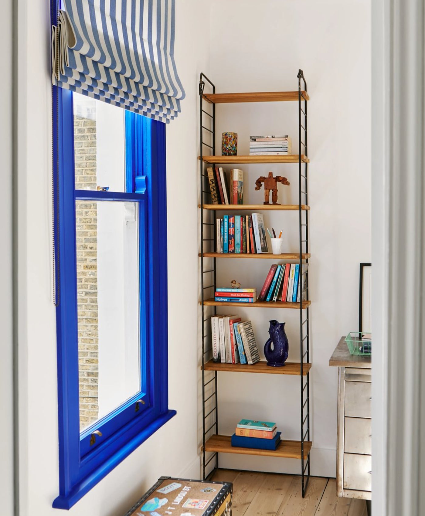 Bookshelf next to a window with blue frame and striped curtain in cool white room