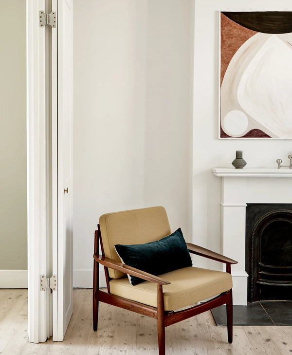 Beige armchair with a dark green pillow in a white room with a fireplace and abstract art.