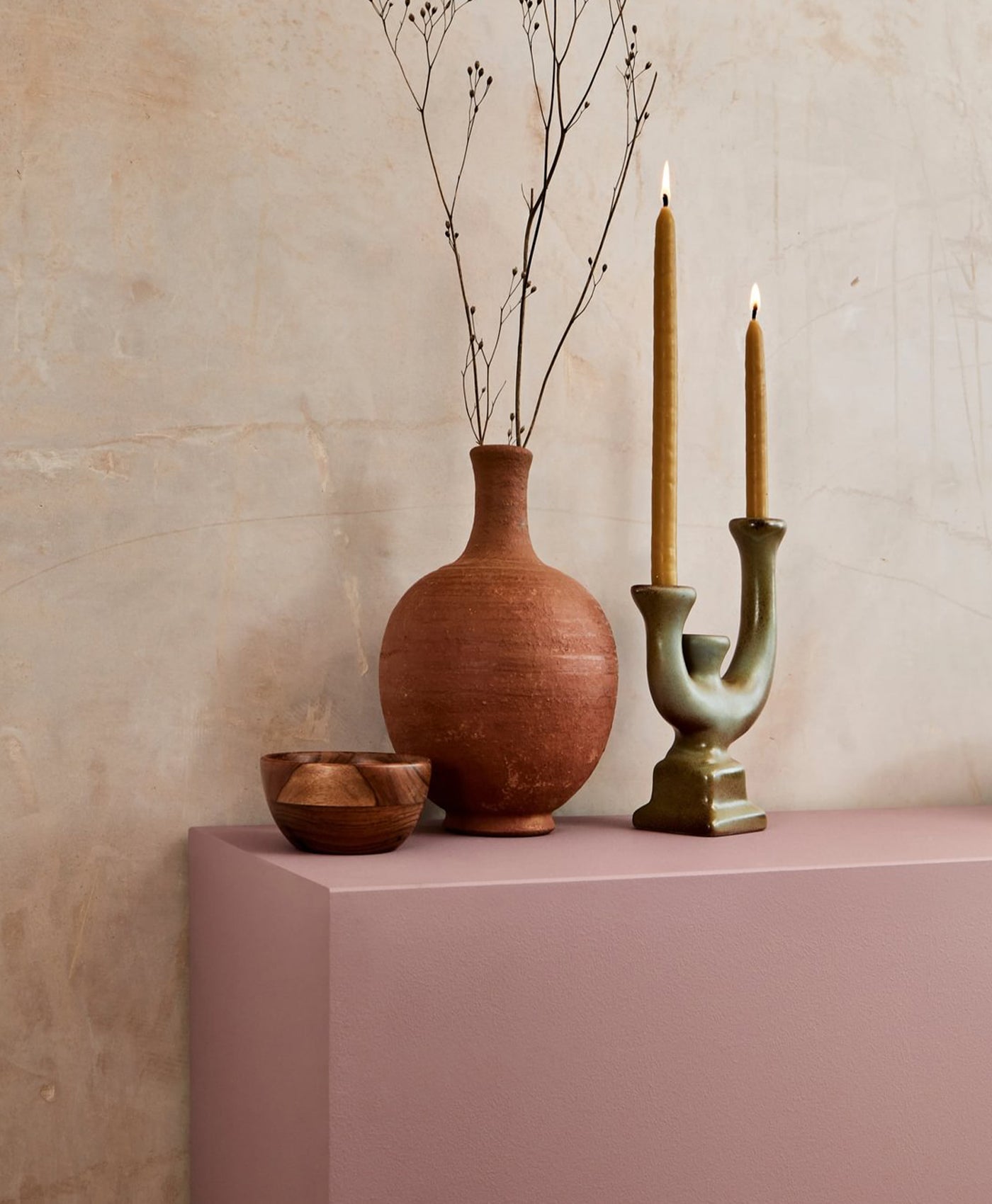 Ceramic vase, bowls, and candle holder on a pink surface with a plaster wall background