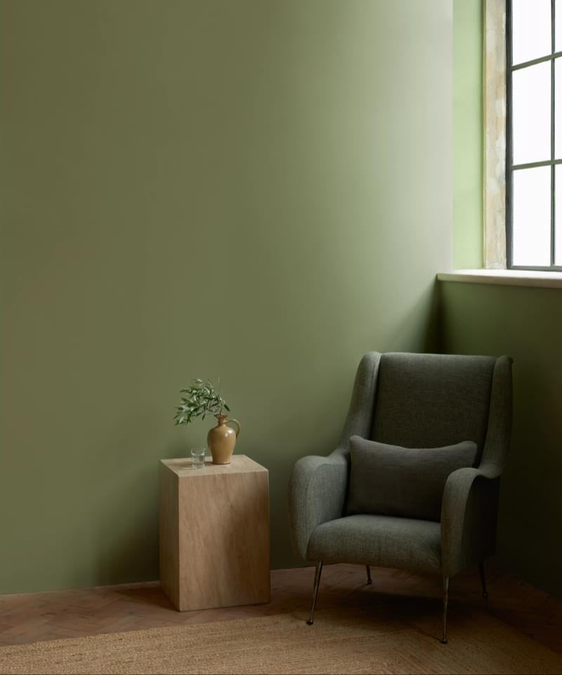 Green armchair in a room with a avocado green wall and a small wooden table with a plant.
